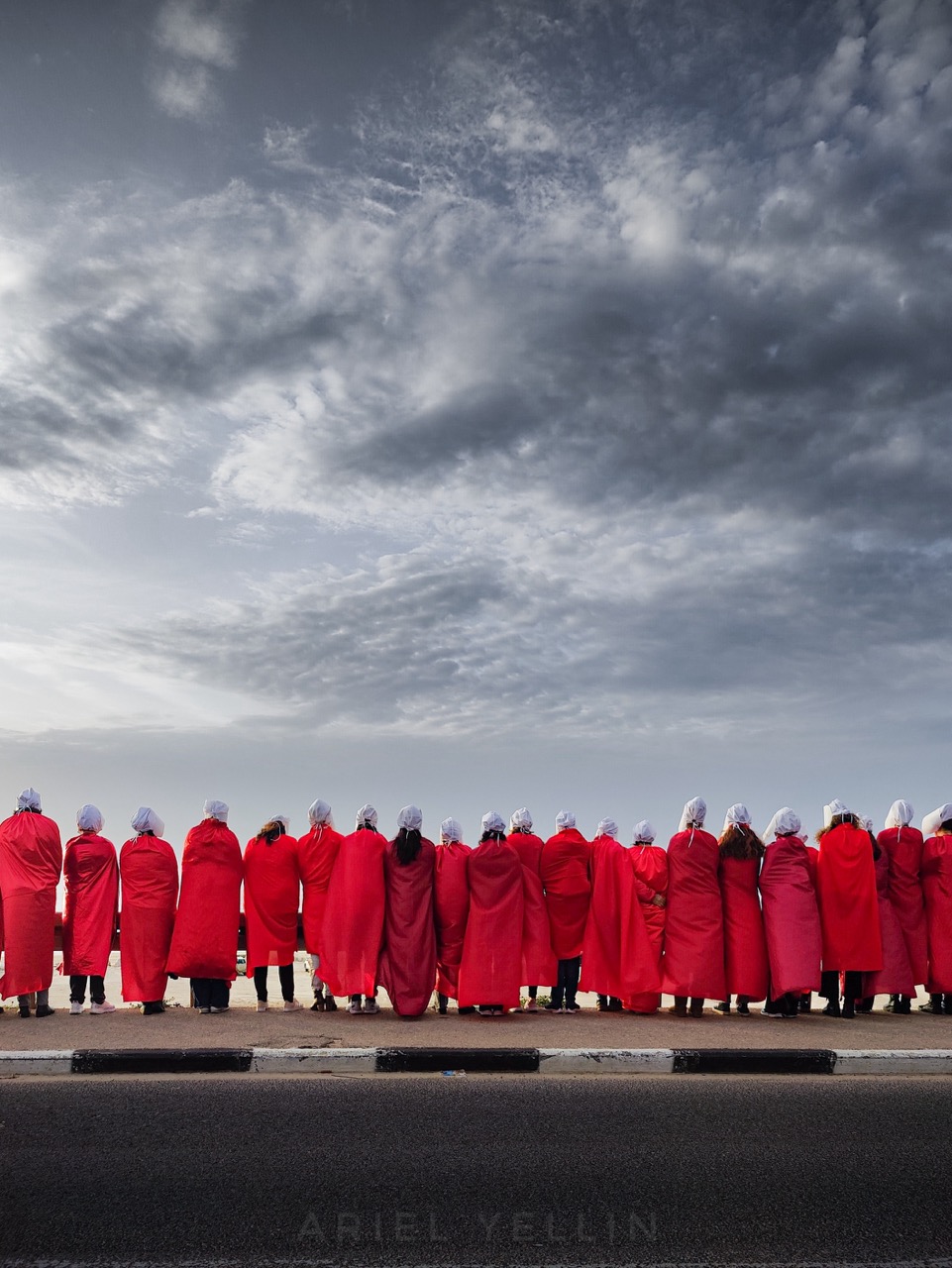 A line of figures in red robes and white bonnets beneath a wide grey sky