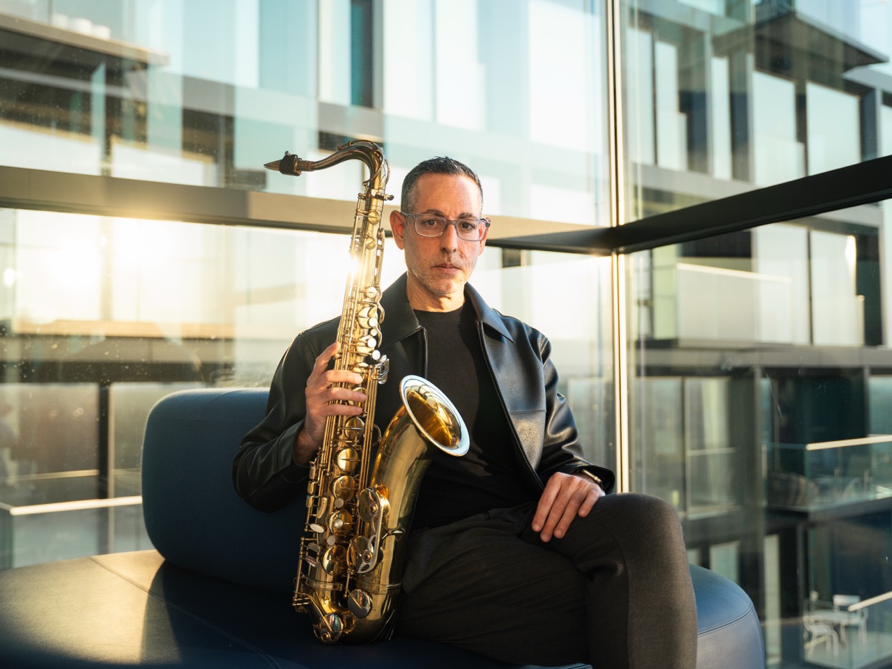 A saxophonist seated beside a glass facade in late light